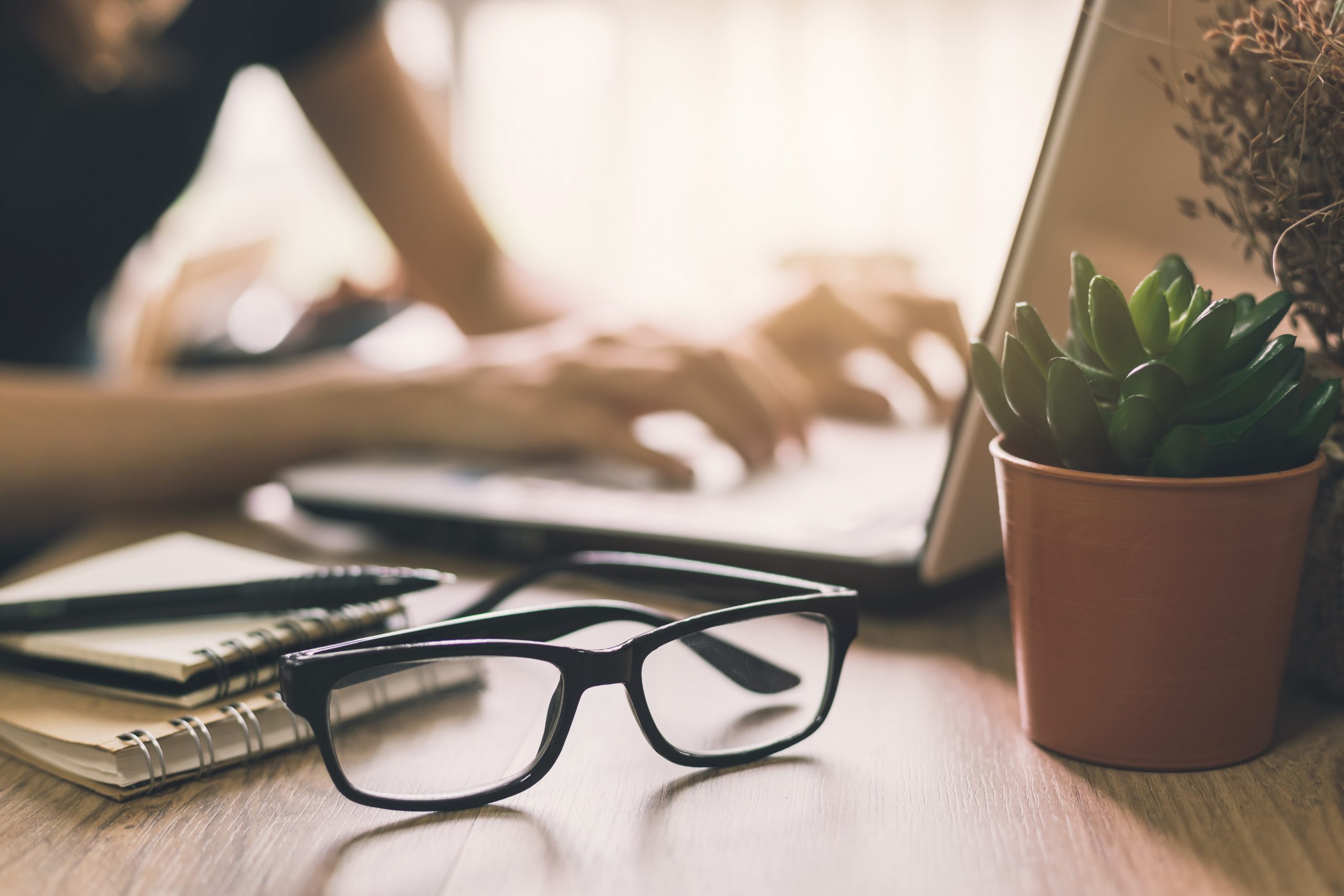 Brown office desk table layout. Notebook, eye-glasses, notebook and plant. Woman sitting in front of computer